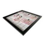 A black photo frame with a grid background, displaying various pieces of jewelry clipped to pink cards using small black clips.