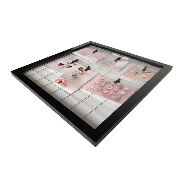 A black photo frame with a grid background, displaying various pieces of jewelry clipped to pink cards using small black clips.