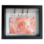 Black photo frame featuring pink roses and a 'You & Me' message on clothespins, creating an elegant and romantic display.