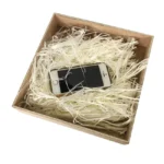 A smartphone resting securely in a box filled with white shredded paper for protective packaging.