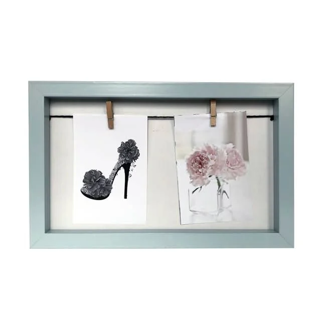 A light blue photo frame displaying two art prints, one of a floral high heel and one of pink flowers in a vase, held by clips.