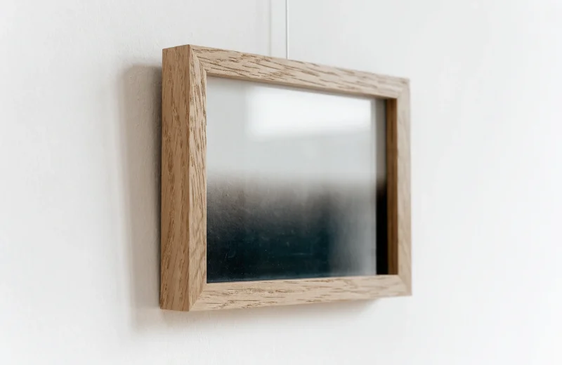 A close-up of a simple, thin photo frame with a light oak finish, hanging on a clean white wall.